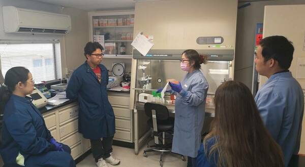 Four lab personnel, including two men and two women, gather around a fifth female lab personnel inside a lab as she explains laboratory procedures.